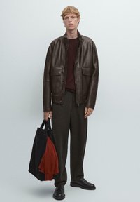 Giacca di pelle marrone con due tasche anteriori, indossata sopra un maglione bordeaux. Pantaloni scuri e una borsa nera con un oggetto rosso all'interno.