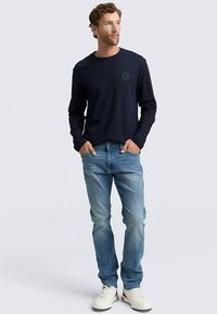 Tamsiai mėlynas marškinėliai ilgomis rankovėmis, suapvalintu kaklu ir krūtinės logotipu, derinami su šviesiai mėlynais džinsais ir baltais sportbačiais. Laisvas stilius kasdieniam dėvėjimui.