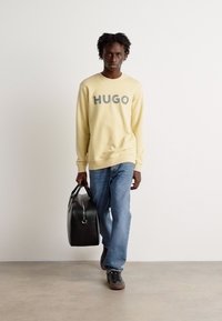 Gul sweatshirt med "HUGO" trykt i grått, parret med blå jeans og sorte sneakers. Modellen bærer en svart treningsbag.