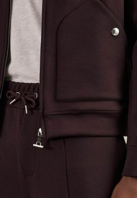 Jachetă sport burgundy cu un buzunar mare, nasture metalic și închidere cu fermoar. Echipată cu pantaloni asortați cu șnur.