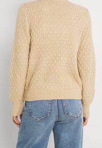 Beiger Kabelstrickpullover mit gerippten Manschetten und Saum, getragen mit hellblauen Jeans, von hinten gezeigt.