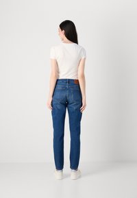 Donna con lunghi capelli scuri, che indossa sneakers bianche, jeans blu e una maglietta bianca a maniche corte, in piedi di spalle contro uno sfondo bianco.