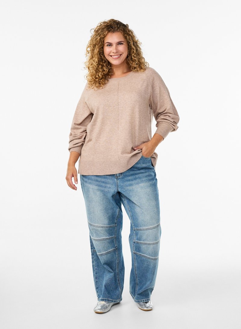 Smilende kvinde med krøllet hår iført en beige sweater, løse blå jeans med knæsyninger og sølvfarvede sko, stående med den ene hånd i lommen.
