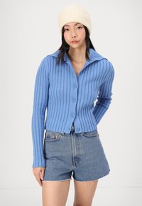 Cardigan bleu clair à côtes avec un col, fermeture à boutons, manches longues, associé à un short en jean dans un délavé bleu.