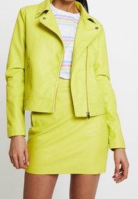 Neonově žlutá kožená motorkářská bunda s asymetrickým zipem, límcem na cvočky a odpovídající přiléhavou mini sukní. Hladká textura, elegantní design.