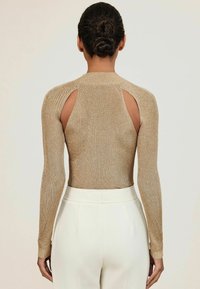 Auksinė ilgomis rankovėmis palaidinė su lygiais raštais, su išpjovimais ant pečių, derinama su baltais aukštai įsegamais kelnais.