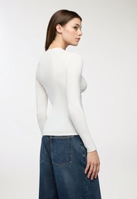 Top lungo a maniche fitte bianco realizzato in un tessuto liscio e elasticizzato, abbinato a jeans in denim blu a vita alta con gamba larga e tasche posteriori.