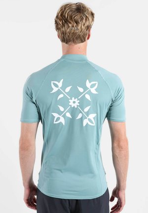 Homme aux cheveux blonds courts portant une chemise bleu clair à manches courtes avec un motif floral blanc comprenant des tiges croisées et des pétales dans le dos.