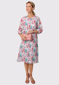 Blumenkleid mit Fältchen, verziert mit pinken und türkisfarbenen Blumen auf einem hellen Hintergrund. Begleitet von einer pinken Clutch und Riemchensandalen.
