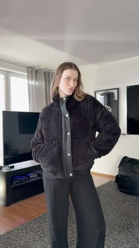 Juodas dirbtinės kailio striukė su spaudėmis, šoninėmis kišenėmis ir apykakle, dėvima virš pilkos mygtukais užsegamos marškinių, derinama su juodomis kelnėmis.