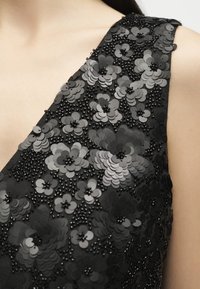 Κοντινή λήψη μαύρου αμάνικου φορέματος με στρωματοποιημένα σχέδια από παγιέτες σε σχήμα λουλουδιών και μικρές διακοσμήσεις με χάντρες στο ύφασμα.