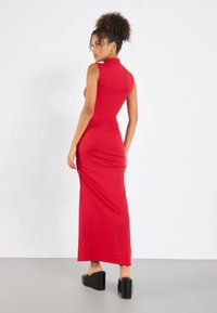 Red ribbed maxi dress with a high neckline and side ruching. Model wears black platform heels, standing on a gray floor.