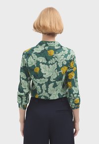Camicia a maniche lunghe con colletto, caratterizzata da un motivo floreale verde e giallo con design fogliosi. Le maniche lunghe sono arrotolate, con una trama di tessuto liscia.