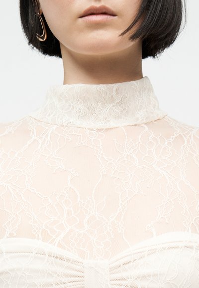 Γυναίκα με διαφανές δαντελένιο τοπ με υψηλό λαιμό και floral σχέδια, με κοντά σκούρα μαλλιά, εμφανίζονται μόνο τα χείλη και το πηγούνι.