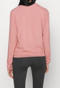 Top a maniche lunghe rosa con scollatura rotonda e orlo arricciato, realizzato in tessuto leggero, abbinato a leggings grigi scuri.