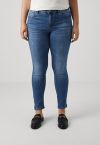 Skinny blue jeans with a faded texture; features five pockets, belt loops, and a zip fly. Worn with black loafers with gold hardware accents.