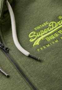 Felpa verde con zip in tessuto texturizzato. Presenta un cappuccio con cordino bianco e un logo ricamato in verde brillante. Chiusura lampo in argento.
