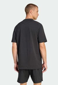 Hombre vestido con una camisa negra de manga corta y suelta y pantalones cortos negros, mostrado de espaldas contra un fondo claro y liso.