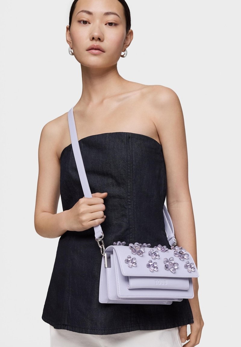 Lilac leather handbag with floral accents, adjustable strap, and a structured shape. Model wears a dark strapless top.