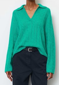 Teal cable-knit sweater with a V-neck and collar, relaxed fit, paired with dark trousers and a black belt. Textured fabric detail evident.