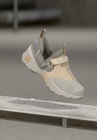 Ανοιχτό μπεζ αθλητικό παπούτσι με υφασμένο επάνω μέρος, εξοπλισμένο με επενδεδυμένο περιλαίμιο, ρυθμιζόμενο λουράκι και λαστιχένια σόλα με λεπτομερή σχέδια πέλματος.