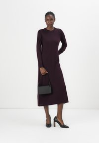 Robe à manches longues, longueur genou, en tricot bordeaux profond. Dotée d'un col rond et d'une silhouette ajustée. Associée à des escarpins noirs et un petit sac à main.