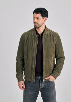 Uomo con giacca bomber in camoscio verde oliva sopra un maglione nero con zip, jeans blu, in piedi davanti a uno sfondo grigio chiaro uniforme.