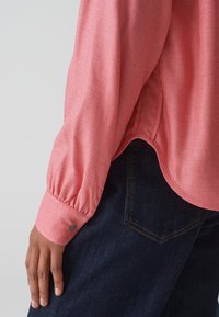 Chemise à manches longues coral en tissu texturé avec des manches bouffantes, dotée d'un poignet boutonné et d'un ourlet arrondi. Associée avec un jean noir.