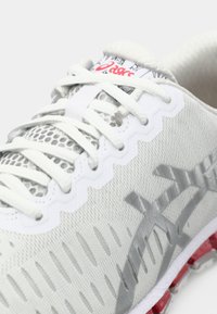 Chaussure de sport gris clair avec des accents en maille, un tissu texturé, des lacets blancs plats et une semelle courbée ornée d'un design rouge et blanc.