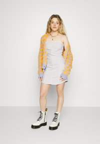 Vestido acinzentado ajustado com alças finas, sobreposto por um cardigan oversized texturizado em laranja e roxo. Botas de combate brancas com atacadores.