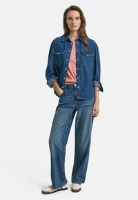 Jeanshemd in Blau, über einem hellen rosa Hemd getragen, kombiniert mit weit geschnittenen blauen Jeans und cremefarbenen Sneakern mit dunklen Akzenten.
