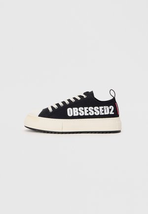 Svarta canvas-sneakers med vit text "OBSESSED2", beige gummisula, platta snören och en dragflik baktill. Sidovy är visad.