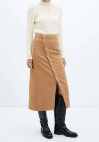 Gonna midi in corduroy color panna con chiusura frontale a bottoni e spacco laterale, abbinata a un maglione in misto lana color crema a trecce e stivali neri al ginocchio.