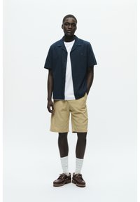 Camicetta navy a maniche corte, t-shirt bianca sotto, pantaloni beige, calze bianche, scarpe marroni con lacci; modello in piedi contro uno sfondo bianco.