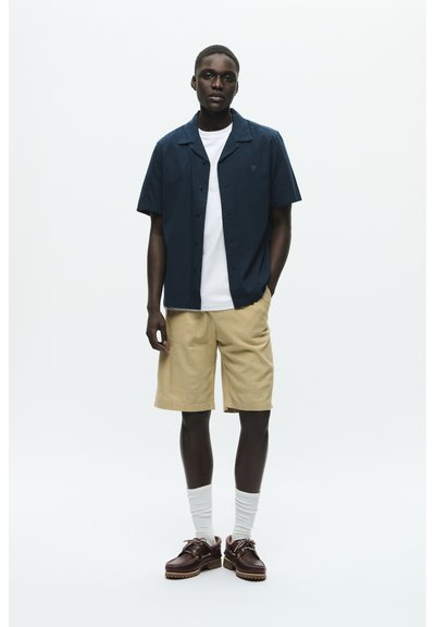 Marineblause mit kurzen Ärmeln, weißes T-Shirt darunter, beige Shorts, weiße Socken, braune Schnürschuhe; Modell steht vor einem weißen Hintergrund.