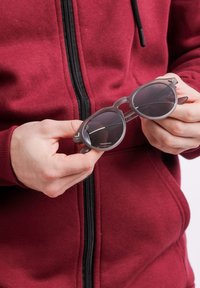 Šedé sluneční brýle se zaobleným tvarem, tmavými skly a texturovaným rámem, které drží osoba oblečená v vínovém bundě na zip.