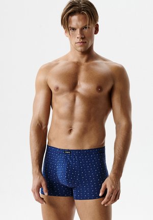 Atletyczny młody mężczyzna z krótkimi włosami, ubrany w niebieskie bokserki z drobnym wzorem białych żaglówek, stojący na jednolitym białym tle.