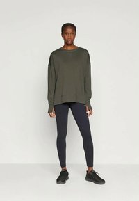 Sudadera de gran tamaño en color verde oliva con mangas largas y escote redondeado, combinada con leggings ajustados en azul marino y zapatillas deportivas negras.