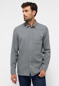 Mann in einem grauen Langarmhemd mit Knopfleiste und Brusttasche sowie dunklen blauen Jeans, der gegen einen weißen Hintergrund nach links blickt.