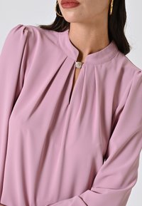 Blusa rosa realizzata in tessuto liscio, con colletto alto, pieghe e un dettaglio a forma di diamante sul collo. Le maniche lunghe offrono una vestibilità sartoriale.