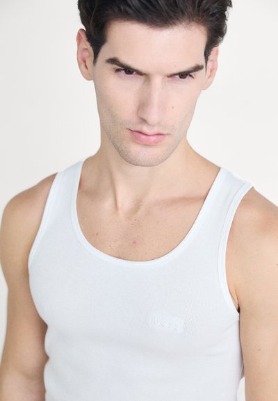 Débardeur en coton blanc avec une texture côtelée et un col rond. Présente un logo embossé discret sur le côté gauche, design ajusté.