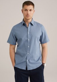 Gaišas zils īsuko piedurkņu, pogājams krekls. Iezīmē klasisku apkakli, redzamu pogu virkni un sarullētas piedurknes.