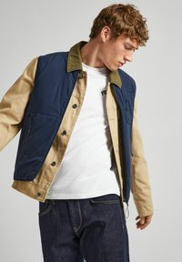 Tvåfärgad jacka med marinblå överdel och beige ärmar. Har tryckknappar, sidofickor och en bomullstextur. Bärs över en vit skjorta.