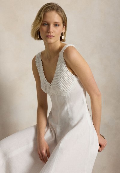 Jauna sieviete ar īsiem blontiem matiem, valkājot baltu kleitu bez piedurknēm ar tamborētu bodiju, sēžot pret vienkāršu bēša fona.