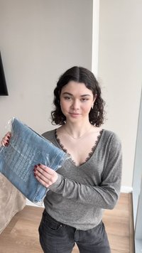 Jeune femme aux cheveux bouclés foncés tenant un jean plié enveloppé dans du plastique, debout à l'intérieur près d'une fenêtre en vêtements décontractés.