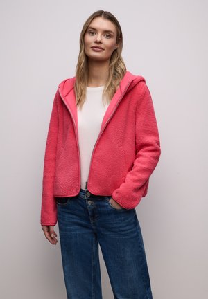 Femme aux longs cheveux blonds portant une veste polaire rose zippée, une chemise blanche et un jean bleu, debout devant un fond clair uni.