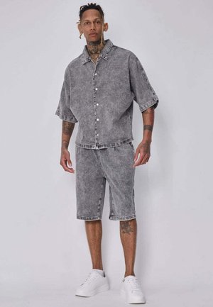 Homme avec des tatouages portant une chemise grise à manches courtes boutonnée et un short gris assorti avec des baskets blanches sur un fond uni.