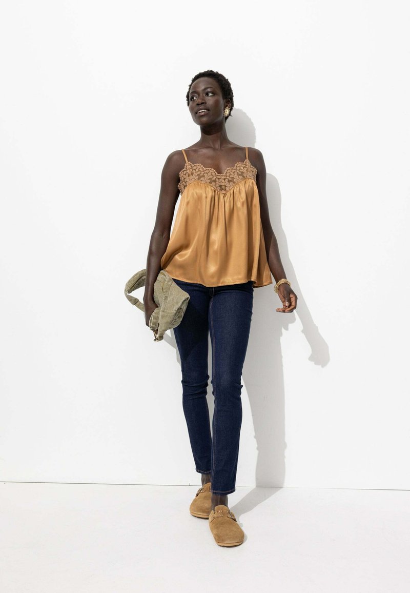 Camisole en satin moutarde avec un bord en dentelle, associé à un jean skinny noir et des mocassins en cuir suédé beige, portant un sac vert olive.