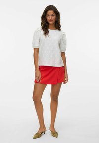 Hvit bluse med puffermer og hullmønster, iført med et rødt miniskjørt. Antrekket er komplementert med lysegrønne spisse flate sko.