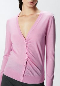 Roza kardigan s dubokim V-izrezom, zatvaranjem na gumbi i nabranom teksturom. Prilagođeni dugi rukavi i rebrasti rub. Glatki pleteni materijal.